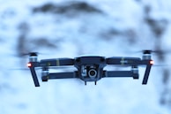A drone is hovering in mid-air against a blurred snowy background. The drone has a sleek design with visible propellers, red lights on either side, and a camera mounted on the front.