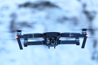 A drone is hovering in mid-air against a blurred snowy background. The drone has a sleek design with visible propellers, red lights on either side, and a camera mounted on the front.