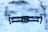 A drone is hovering in mid-air against a blurred snowy background. The drone has a sleek design with visible propellers, red lights on either side, and a camera mounted on the front.