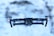 A drone is hovering in mid-air against a blurred snowy background. The drone has a sleek design with visible propellers, red lights on either side, and a camera mounted on the front.