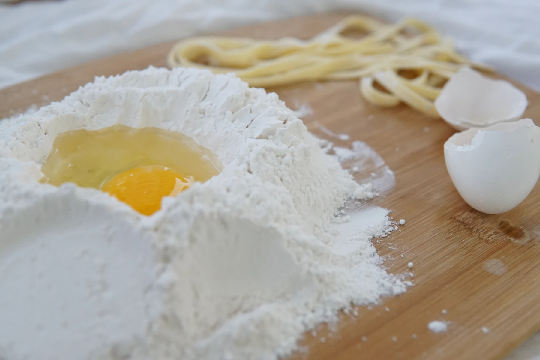 An egg yolk sitting in a well of flour on a wooden board, with cut noodles and cracked shells behind