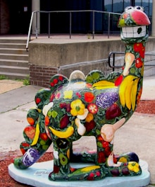 A whimsical, cartoonish dinosaur sculpture is painted with vibrant colors and decorated with various fruits and vegetables, creating a playful and creative design. The dinosaur has big, cheerful eyes and a broad smile, with its surface covered in bananas, peppers, cabbage, and flowers among other items.