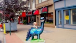 A colorful dinosaur sculpture stands on a city sidewalk, with a young child figure riding on its back. Surrounding the installation are various storefronts with red awnings and signs. The street is lined with trees, and a couple of pedestrians are visible in the distance.