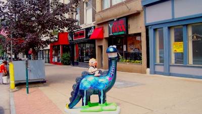 A colorful dinosaur sculpture stands on a city sidewalk, with a young child figure riding on its back. Surrounding the installation are various storefronts with red awnings and signs. The street is lined with trees, and a couple of pedestrians are visible in the distance.