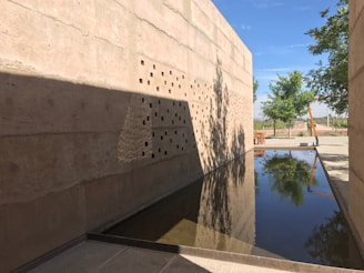 A modern architectural space with tall concrete walls featuring small square cutouts. A narrow, reflective pool runs along the ground, capturing the reflections of trees and the clear blue sky. Shadows and patterns are cast on the walls, adding texture and depth. In the background, there are trees and open space, suggesting an outdoor setting.