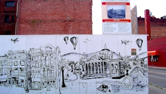 A city street art mural depicts historical buildings with intricate, detailed lines. Above the mural, a signboard advertises the development of the area. The mural features hot air balloons, airplanes, and a map-like design of roads and trees.
