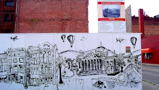 A city street art mural depicts historical buildings with intricate, detailed lines. Above the mural, a signboard advertises the development of the area. The mural features hot air balloons, airplanes, and a map-like design of roads and trees.