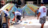 Several people are collaboratively working on a vibrant outdoor mosaic art project. The mosaic features a wide array of colorful tiles forming an intricate pattern, mounted on a curved wall. The participants are using tools and materials like buckets, paint, and brushes, indicating an active creation process. The scene suggests a community or group effort to beautify a public space.
