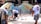 Several people are collaboratively working on a vibrant outdoor mosaic art project. The mosaic features a wide array of colorful tiles forming an intricate pattern, mounted on a curved wall. The participants are using tools and materials like buckets, paint, and brushes, indicating an active creation process. The scene suggests a community or group effort to beautify a public space.