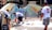 Several people are collaboratively working on a vibrant outdoor mosaic art project. The mosaic features a wide array of colorful tiles forming an intricate pattern, mounted on a curved wall. The participants are using tools and materials like buckets, paint, and brushes, indicating an active creation process. The scene suggests a community or group effort to beautify a public space.