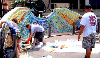 Several people are collaboratively working on a vibrant outdoor mosaic art project. The mosaic features a wide array of colorful tiles forming an intricate pattern, mounted on a curved wall. The participants are using tools and materials like buckets, paint, and brushes, indicating an active creation process. The scene suggests a community or group effort to beautify a public space.