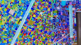 A vibrant mosaic artwork composed of multiple small, colorful tiles arranged in a dynamic, abstract pattern. The tiles vary in color and size, creating an intricate and lively visual effect.