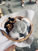 A hand holding a bowl made from a coconut shell, filled with scoops of ice cream topped with jelly-like black cubes, small round seeds, and roasted peanuts. The bowl rests on a paper napkin, and the background shows a paved surface with a few feet visible.