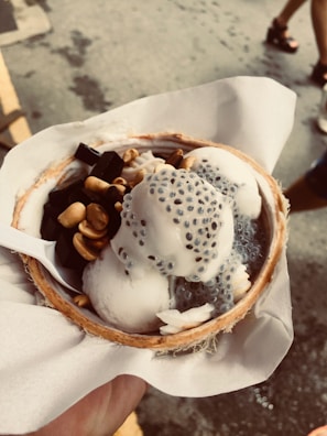 A bowl made from half a coconut contains scoops of ice cream topped with chia seeds, pieces of grass jelly, and peanuts. The bowl is held above a textured gray surface, and a white paper napkin is used as a holder.