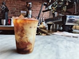 A chilled coffee drink with swirls of cream sits in a plastic cup on a marble countertop. The background features a rustic brick wall, various coffee-making equipment, and an espresso machine. The atmosphere suggests a cozy, urban coffee shop.