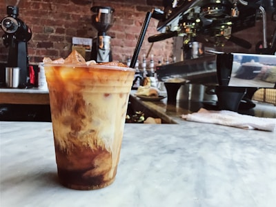 A chilled coffee drink with swirls of cream sits in a plastic cup on a marble countertop. The background features a rustic brick wall, various coffee-making equipment, and an espresso machine. The atmosphere suggests a cozy, urban coffee shop.