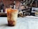 A chilled coffee drink with swirls of cream sits in a plastic cup on a marble countertop. The background features a rustic brick wall, various coffee-making equipment, and an espresso machine. The atmosphere suggests a cozy, urban coffee shop.