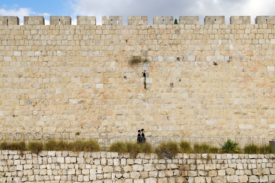 two person walking beside beige brick wall,