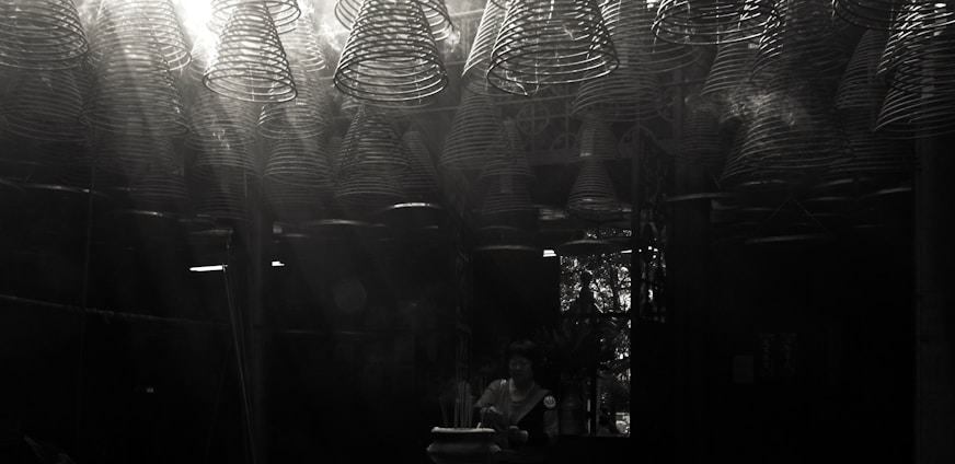 A dimly lit space filled with intricate, spiral incense coils hanging from the ceiling, casting light patterns. A person stands beneath, engaging in a meditative or spiritual activity, with subtle light streaming through a window in the background.