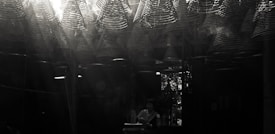 A dimly lit space filled with intricate, spiral incense coils hanging from the ceiling, casting light patterns. A person stands beneath, engaging in a meditative or spiritual activity, with subtle light streaming through a window in the background.