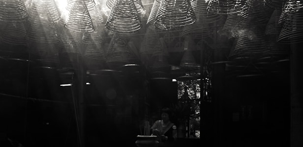 A dimly lit space filled with intricate, spiral incense coils hanging from the ceiling, casting light patterns. A person stands beneath, engaging in a meditative or spiritual activity, with subtle light streaming through a window in the background.