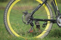 A close-up view of a mountain bike's rear wheel showing the gear mechanism, chain, and part of the frame. The bicycle tire has a yellow rim, and the frame is black. The bike is resting on green grass, suggesting an outdoor setting.