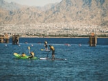 People engage in water sports on a calm body of water with mountainous terrain in the background. Some individuals are kayaking while others are paddleboarding. Brightly colored life jackets and paddles are prominent, with the urban backdrop adding contrast between nature and city life.