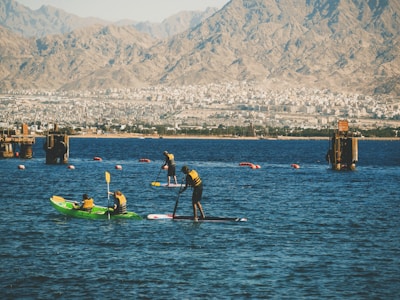 People engage in water sports on a calm body of water with mountainous terrain in the background. Some individuals are kayaking while others are paddleboarding. Brightly colored life jackets and paddles are prominent, with the urban backdrop adding contrast between nature and city life.