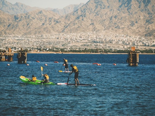 People engage in water sports on a calm body of water with mountainous terrain in the background. Some individuals are kayaking while others are paddleboarding. Brightly colored life jackets and paddles are prominent, with the urban backdrop adding contrast between nature and city life.