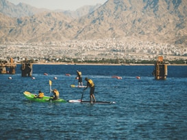 People engage in water sports on a calm body of water with mountainous terrain in the background. Some individuals are kayaking while others are paddleboarding. Brightly colored life jackets and paddles are prominent, with the urban backdrop adding contrast between nature and city life.