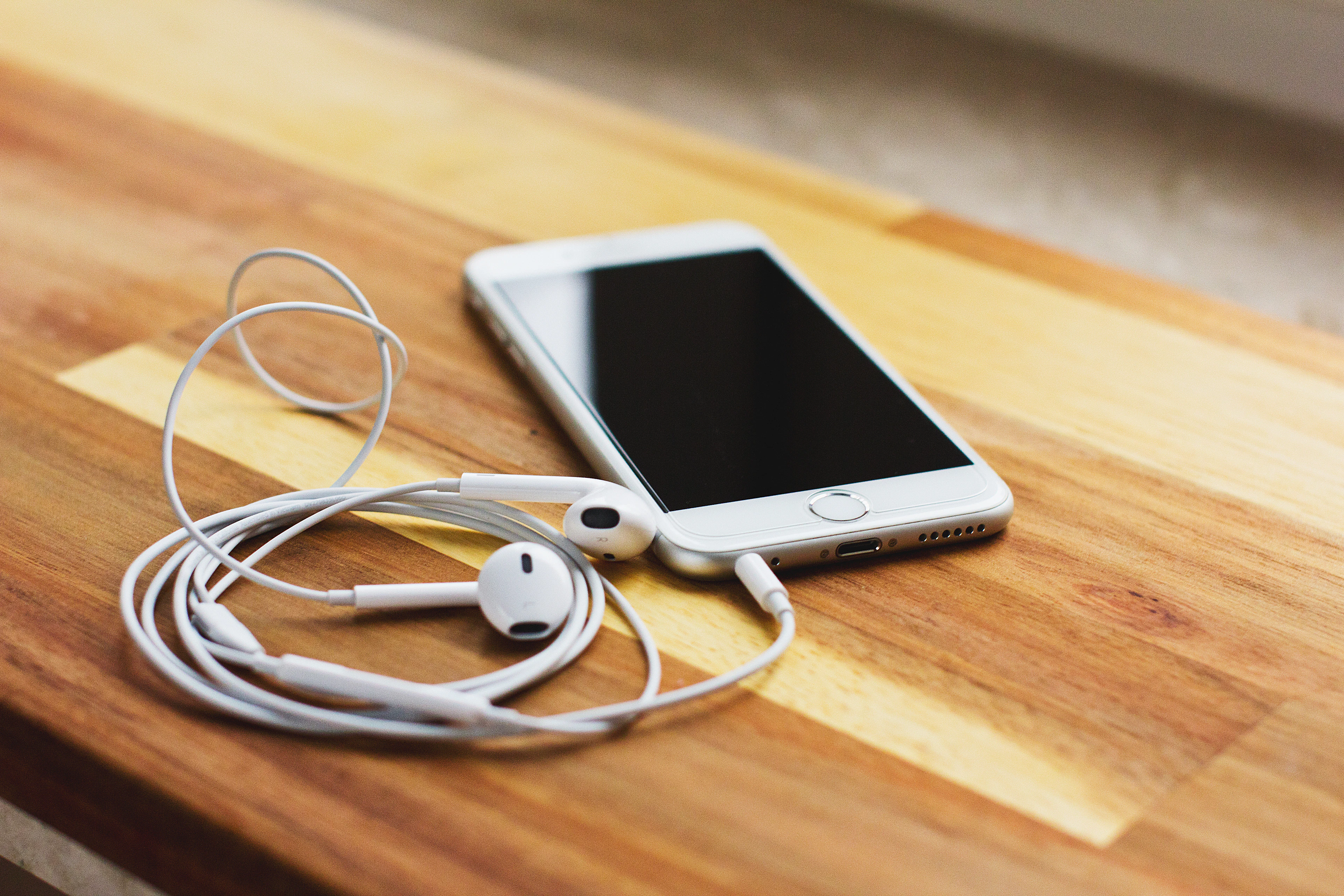یک iPhone 6 نقره‌ای با EarPods سیمی سفید روی میز چوبی قهوه‌ای