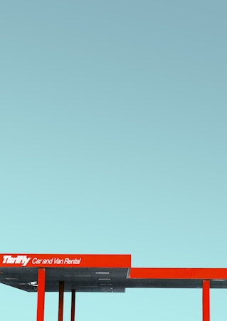 A minimalist photograph featuring a red and grey canopy with the sign 'Car and Van Rental' against a clear blue sky. The structure has a modern and geometric design.