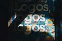 The image features a vibrant collage of logos and graphic designs on a dark background. Bold white text reading 'Logos, logos, logos' is prominently displayed over the colorful backdrop. Various symbols and shapes, including sun-like designs and abstract patterns, fill the scene, creating an artistic and energetic composition.