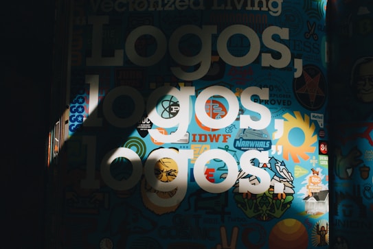 The image features a vibrant collage of logos and graphic designs on a dark background. Bold white text reading 'Logos, logos, logos' is prominently displayed over the colorful backdrop. Various symbols and shapes, including sun-like designs and abstract patterns, fill the scene, creating an artistic and energetic composition.
