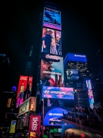 A lively scene in a busy urban area featuring a large number of illuminated billboards and advertisements. Various brands like Coca-Cola, Hyundai, and Broadway shows are prominently displayed. A crowd of people is gathered around, adding to the bustling atmosphere.