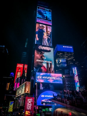 A lively scene in a busy urban area featuring a large number of illuminated billboards and advertisements. Various brands like Coca-Cola, Hyundai, and Broadway shows are prominently displayed. A crowd of people is gathered around, adding to the bustling atmosphere.