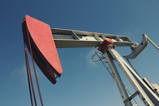 A large metal oil pumpjack with a red rotary head is set against a clear blue sky. The structure includes metal beams, cables, and a ladder. The machinery shows signs of wear with visible rust and peeling paint.