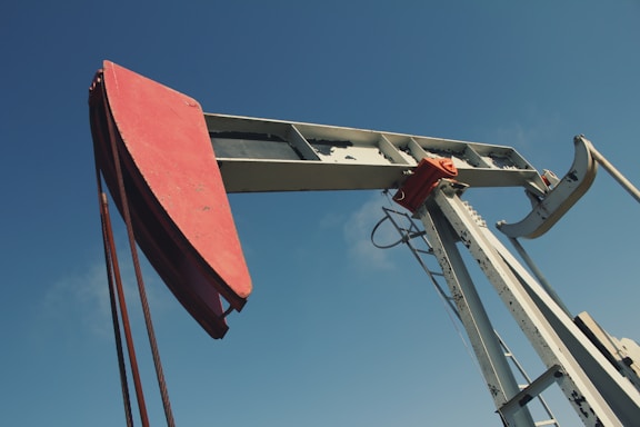 A large metal oil pumpjack with a red rotary head is set against a clear blue sky. The structure includes metal beams, cables, and a ladder. The machinery shows signs of wear with visible rust and peeling paint.