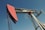 A large metal oil pumpjack with a red rotary head is set against a clear blue sky. The structure includes metal beams, cables, and a ladder. The machinery shows signs of wear with visible rust and peeling paint.