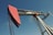 A large metal oil pumpjack with a red rotary head is set against a clear blue sky. The structure includes metal beams, cables, and a ladder. The machinery shows signs of wear with visible rust and peeling paint.