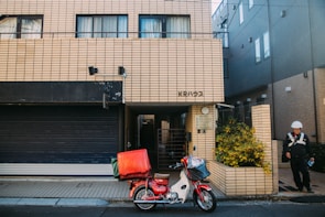 A brick building with a motorbike parked in front. The motorbike has a large red and green delivery box attached. There is a person in uniform and a helmet standing on the sidewalk next to the building. The entrance to the building is surrounded by small plants.