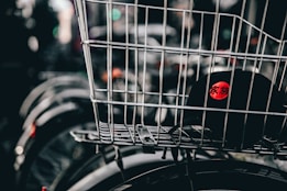 A wire basket is mounted on a bicycle, containing a black cap featuring a red circle with characters printed in white. The image focuses primarily on the basket and cap, while the background is blurred, showing elements like other bicycle wheels.