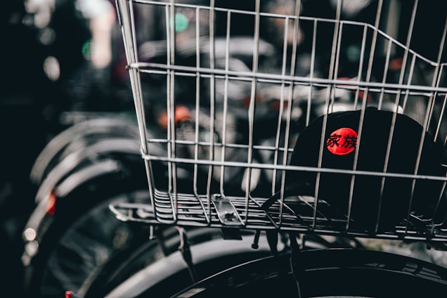 A wire basket is mounted on a bicycle, containing a black cap featuring a red circle with characters printed in white. The image focuses primarily on the basket and cap, while the background is blurred, showing elements like other bicycle wheels.