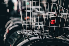 A wire basket is mounted on a bicycle, containing a black cap featuring a red circle with characters printed in white. The image focuses primarily on the basket and cap, while the background is blurred, showing elements like other bicycle wheels.