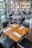A modern restaurant interior with neatly arranged wooden tables, each set with glasses, white napkins, and cutlery. Large windows provide ample natural light and offer a view of the surrounding cityscape with brick and glass buildings.