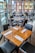 A modern restaurant interior with neatly arranged wooden tables, each set with glasses, white napkins, and cutlery. Large windows provide ample natural light and offer a view of the surrounding cityscape with brick and glass buildings.