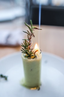A unique candle featuring an herb sprig, possibly rosemary, protruding from its top. The flame is small and emits a thin trail of smoke. The candle is placed on a white plate.