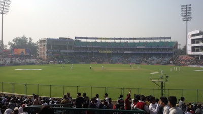 A cricket stadium filled with spectators, with players on the field wearing white uniforms. The stands are populated, and large floodlights are visible. Advertisements are displayed around the stadium, and a bright digital screen is on the left side.