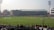 A cricket stadium filled with spectators, with players on the field wearing white uniforms. The stands are populated, and large floodlights are visible. Advertisements are displayed around the stadium, and a bright digital screen is on the left side.