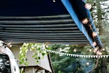 A striped blue awning extends over a scene decorated with string lights. A trailer with a reflective surface is partially visible, alongside some leafy branches and a building. A forest background adds to the outdoor atmosphere.